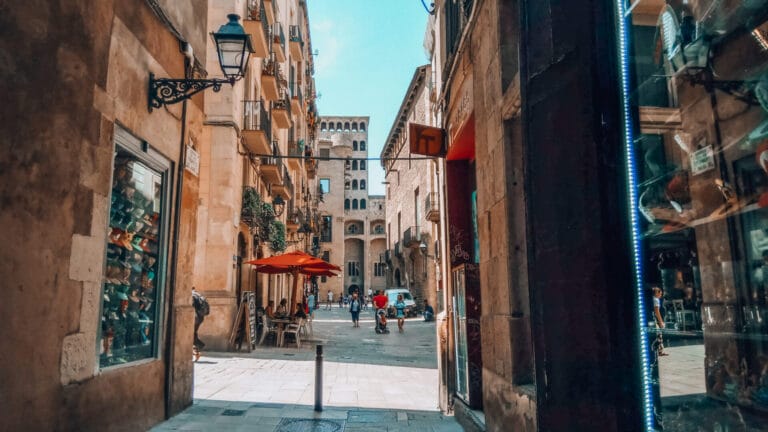View from Carrer del Veguer toward Plaça del Rei in Barcelona’s Gothic Quarter, with people, cafés, and medieval architecture.