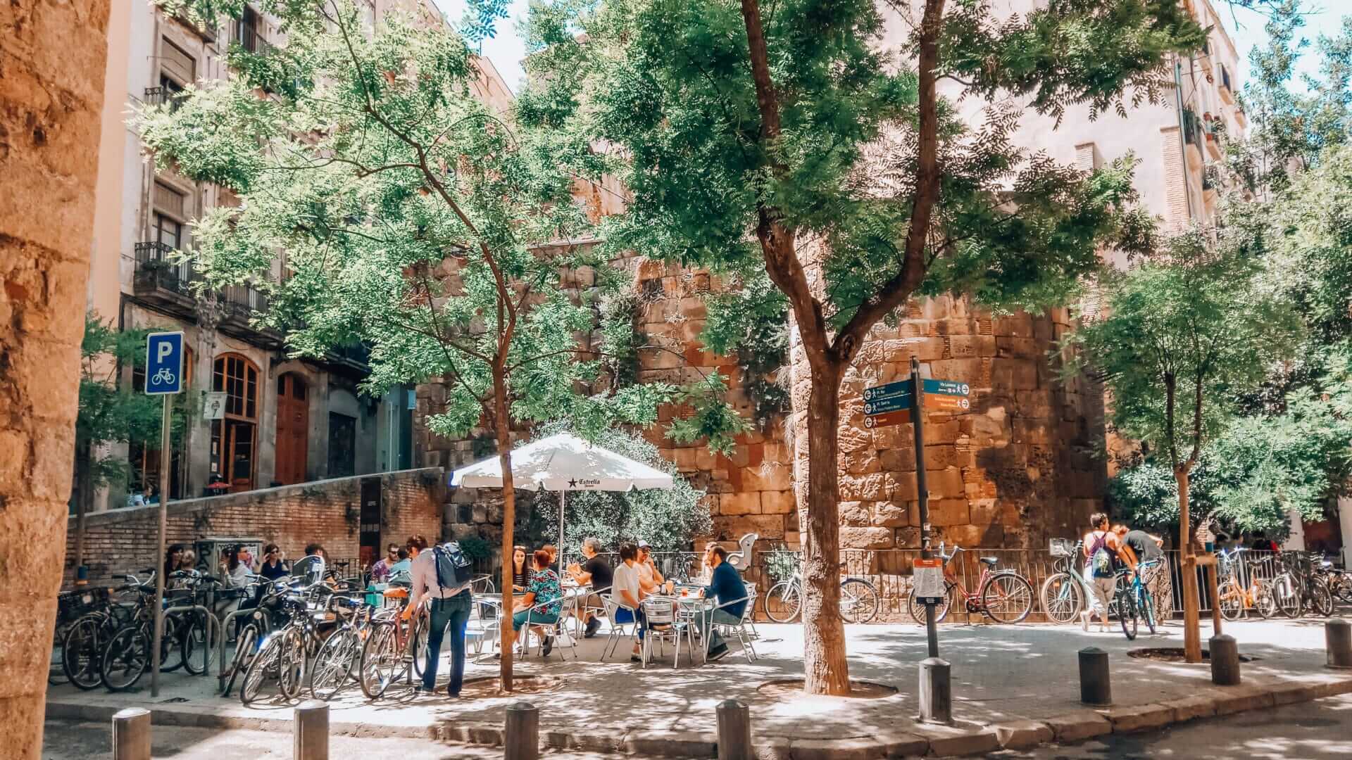 Plaça dels Traginers in Barcelona's Gothic Quarter with people at an outdoor café, bicycles, and a well-preserved Roman wall.